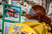 Woman while hiking with snowshoes on her backpack reads bus timetable, Spitzing area, Bavarian Alps, Upper Bavaria, Bavaria, Germany