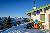Man and woman hiking with snowshoes on their rucksacks sit on bench in front of snow-covered hut and drink tea, Enzianhütte, Hochgern, Chiemgau Alps, Upper Bavaria, Bavaria, Germany