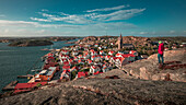Mann blickt über Küste und Dorf Fjällbacka vom Berg Vetteberget mit Felsspalte von oben bei Tag mit Sonne und blauem Himmel an der Westküste in Schweden\n
