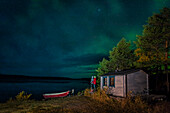 Paar beobachtet Polarlichter im Nachthimmel an Hütte am Seeufer mit Booten in Lappland, Schweden\n