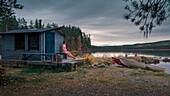 Frau sitzt vor Sauna am See Stor-Skabram in Lappland, Schweden