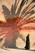 Mann in Roben zu Fuß durch die Wand mit Palmenschatten, Essaouira, Marokko