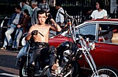 Shirtless man wearing leather pants with smoking a cigarette and combing his hair while sitting on a motorcycle in a busy street, Tokyo, Japan