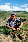 Porträt eines älteren chinesischen Bauern, der Pflanzen hält, während er auf einer Weide sitzt, China