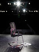 Rückansicht eines punktbeleuchteten leeren Stuhls auf der Bühne mit einem Skript und einem Glas Wasser daneben und Blick auf einen leeren Zuschauerraum