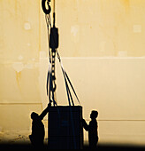 Silhouette of two men guiding cargo attached to large hook in front of ships hull