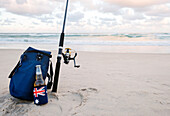 Bier im Stubbie-Halter mit australischer Flagge, Kühltasche und Angelrute im Sand am Strand
