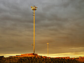 Tall floodlights shining on export log pile with stormy early evening sky