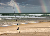 Surf Caster Angelrute im Sand gesichert und doppelter Regenbogen im stürmischen Himmel am Strand