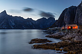 Rote Hütten beleuchtet am Fjord vor Bergkulisse. Hamnoy, Reine, Lofoten, Nordland, Norwegen.