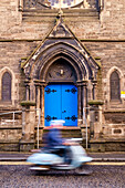 Person auf einem Roller, der an einem gotischen Bogen mit hellblauer Tür vorbeifährt, Edinburgh, UK