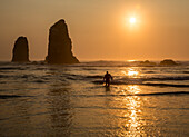 Surfer walking out of the sea with sun setting behind rock in waves on beach.