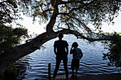Zwei Kinder am Rand einer Wasserstraße, Rückansicht nebeneinander, im Okavango Delta, Botswana.