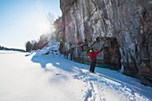 A man in a red jacket standing in front of an ice cliff looking up, ice climbing.