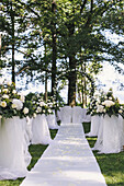 A garden with tables laid under the shade of tall trees, a floral arch, setting for a wedding