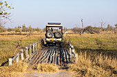 Ein Safarifahrzeug, das eine Brücke in der Landschaft von Sümpfen und Wasserstraßen überquert, Okavango-Delta, Botswana