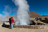 Touristen, die den Dampf betrachten, der aus heißen Quellen im geothermischen Becken der Geysire El Tatio in der Nähe von San Pedro de Atacama in der Atacama-Wüste im Norden Chiles aufsteigt.