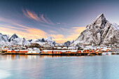 Wintersonnenuntergang über den schneebedeckten Bergen und dem Dorf Sakrisoy am gefrorenen Meer, Reine, Nordland, Lofoten, Norwegen, Skandinavien, Europa
