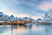Holzhütten der Fischer, die bei Sonnenuntergang in dem winzigen Dorf Sakrisoy, Reine, Nordland, Lofoten-Inseln, Norwegen, Skandinavien, Europa mit Schnee bedeckt sind