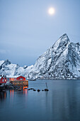 Rote Rorbu-Hütten und majestätischer Olstind-Gipfel, der während der Winterdämmerung vom Mond beleuchtet wird, Hamnoy, Nordland County, Lofoten-Inseln, Norwegen, Skandinavien, Europa
