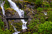 Ein Mann auf einer Fußgängerbrücke überquert einen der vielen Wasserfälle, die ihren Ursprung am Briksdal-Gletscher, Stryn, Vestland, Norwegen, Skandinavien, Europa haben