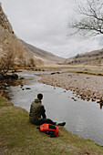 Männlicher Wanderer rastet an einem ruhigen Bach in abgelegener Landschaft, Glen Nevis, Schottische Highlands, Schottland