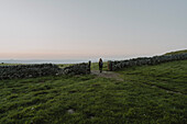 Woman hiking through stone wall gate in rural landscape at sunrise, Castleton, Derbyshire, England