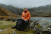 Männlicher Wanderer ruht auf Felsen am Bergsee Llyn Padarn, Snowdonia, Wales