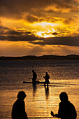 Silhouetted family with paddleboard in ocean below dramatic sky at sunset