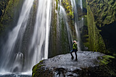Iceland, Seljalandsfoss, woman standing on large rock in front of streaming falls in green slot canyon. (MR)