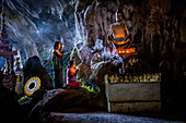 A cave temple with Buddha statues, lit candles and two girls kneeling and praying.