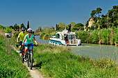Zwei Personen fahren am Canal du Midi Rad, Boot im Hintergrund, bei Capestang, Canal du Midi, UNESCO Welterbe Canal du Midi, Okzitanien, Frankreich