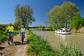 Zwei Personen fahren am Canal du Midi Rad, Boot im Hintergrund, bei Capestang, Canal du Midi, UNESCO Welterbe Canal du Midi, Okzitanien, Frankreich