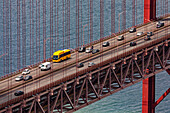 Detail der Brücke des 25. April über den Tejo mit Autos und einem gelben Bus unter den Brückenpfeilern, Lissabon, Portugal