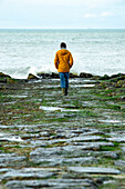 Woman in orange jacket and blue pants walking towards the sea.