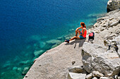 Woman hiking sitting on rock slab over mountain lake, Estany de Mar, Aigüestortes i Estany de Sant Maurici National Park, Catalonia, Pyrenees, Spain