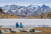 Man and woman sitting at Lej Lunghin, Piz Bernina and Piz Roseg in the background, Lunghinsee, Innquelle, Albula Alps, Graubünden, Switzerland