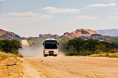 A modern bus for tourists drives on a dusty gravel road through the rocks in the Erongo Mountains, Namibia, Africa
