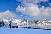 Verschneites rotes Haus mit Fjord und Berge im Hintergrund, Senjahopen, Senja, Troms og Finnmark, Norwegen