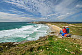 Mann und Frau beim Wandern sitzen auf Felsen und blicken auf den Strand Plage de Pen Hat, Camaret-sur-Mer, GR 34, Zöllnerweg, Sentier Côtier, Halbinsel Crozon, Atlantikküste, Bretagne, Frankreich