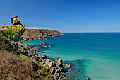 Frau beim Wandern sitzt auf Felsvorsprung und blickt aufs Meer, Cap-Sizun, GR 34, Zöllnerweg, Bretagne, Frankreich