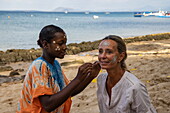 Einheimische Frau trägt Masonjoany Gesichtsbemalung von  auf eine blonde Besucherin auf, Nosy Komba, Diana, Madagaskar, Indischer Ozean