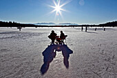 Pärchen sitzt auf Klappstühlen auf zugefrorenem See neben Fußgänger und Eisläufer, Südtirol, Trentino, Italien