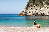  Frau sitzt auf einem Felsen am Strand der Dafines-Bucht, Halbinsel Karaburun, im Karaburun-Sazan-Meerespark, Bucht von Vlore, Albanien, Südosteuropa 