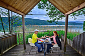  Man and woman sitting on the terrace of a tiny house and toasting each other, reed bivouac, Via paradiso, Millstätter See, Nockberge, Niedere Tauern, Carinthia, Austria 