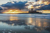  La Rocco Tower stands on the sandy beach at sunset. The sky is reflected in the water. 