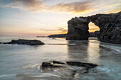  Rock arch on the beach of Playa de las Catedralas. Wave movement and small rock in the water in the foreground. Golden hour. 