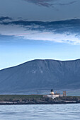  Graemsey low lighthouse in the evening light in front of mountains. Calm sea in the foreground. 