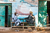 Elderly men sitting on a bench under a mural in a street of the village of Ramadi, west bank of the Nile south of Edfu, Egypt, North East Africa