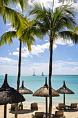  Thatched umbrellas on the beach with coconut trees at Royal Palms Beachcomber Luxury (Beachcomber Resorts) and catamaran sailboats in the distance, Grand Baie, Rivière du Rempart, Mauritius, Indian Ocean 